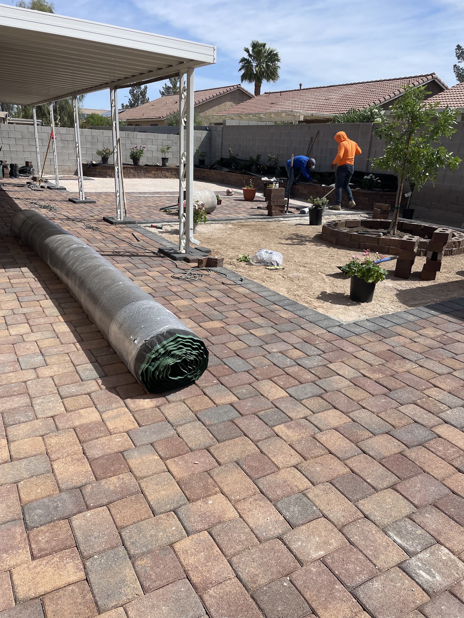 Backyard paver patio installation in progress with turf underlayment roll and workers building a planter